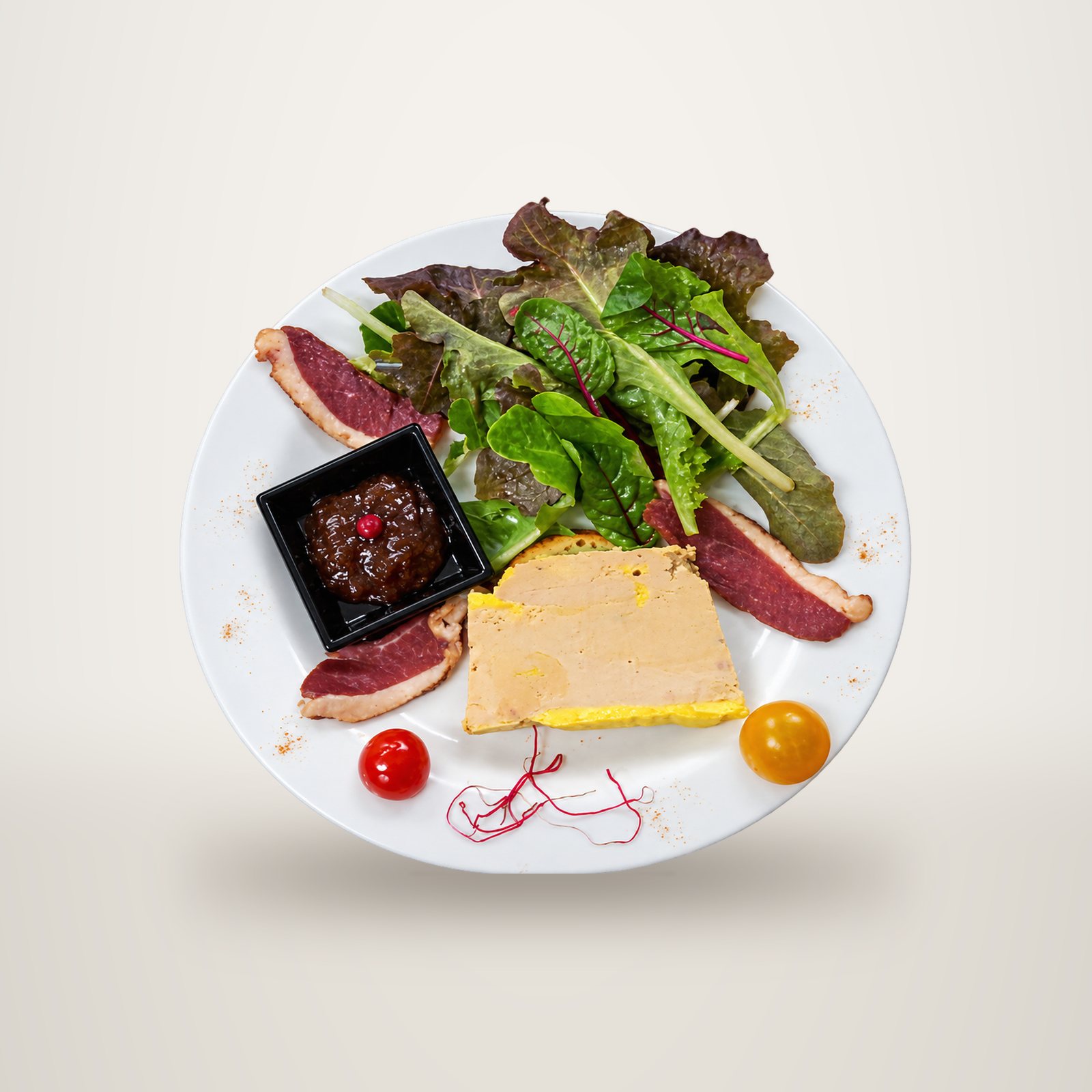 Vue de dessus d'une assiette blanche garnie d'une tranche de foie gras, de lamelles de magret séché, de salade, de tomates cerises et d'un petit pot de chutney, posée sur un plan de travail en inox.