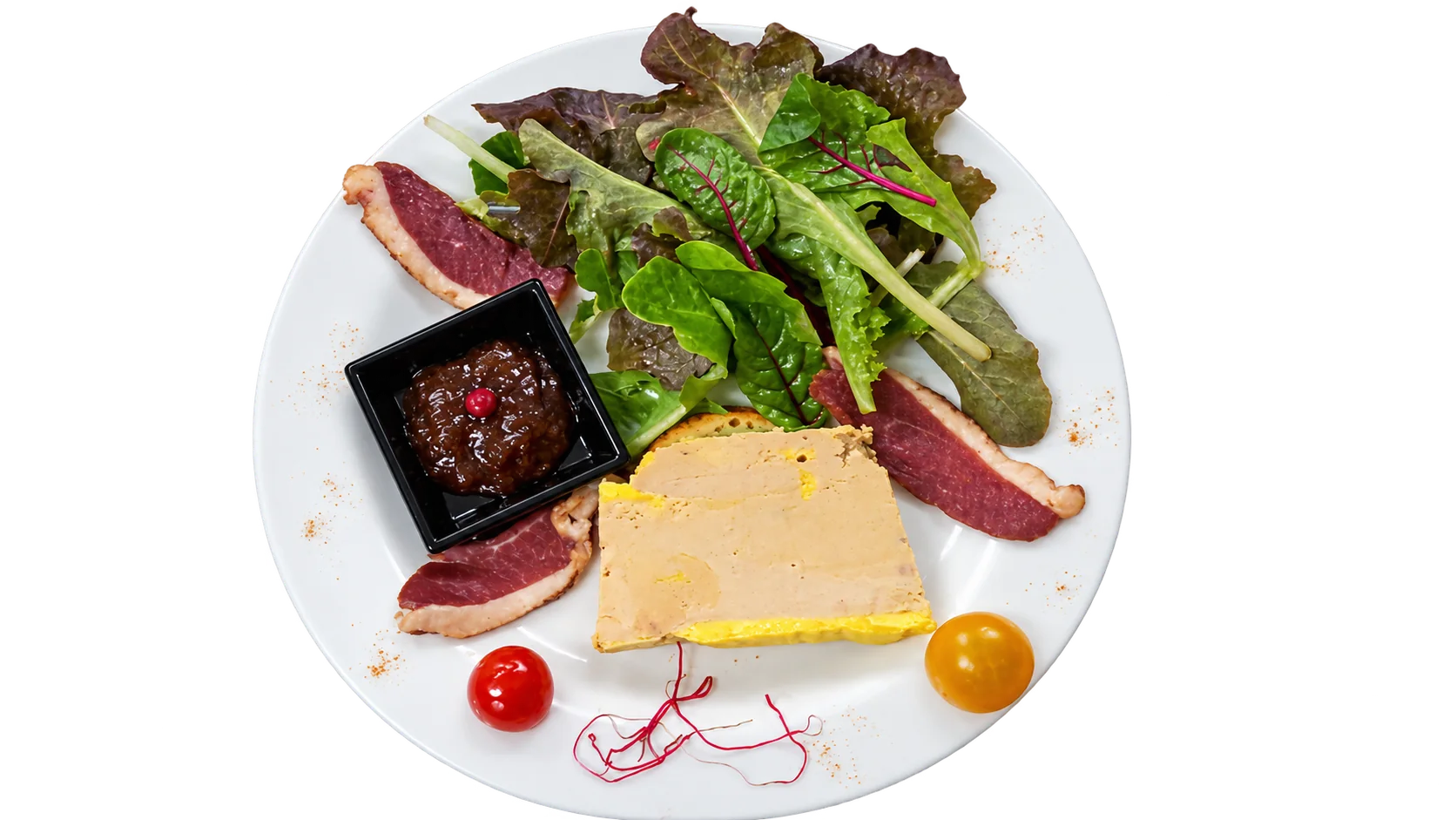 Vue de dessus d'une assiette blanche garnie d'une tranche de foie gras, de lamelles de magret séché, de salade, de tomates cerises et d'un petit pot de chutney, posée sur un plan de travail en inox.
