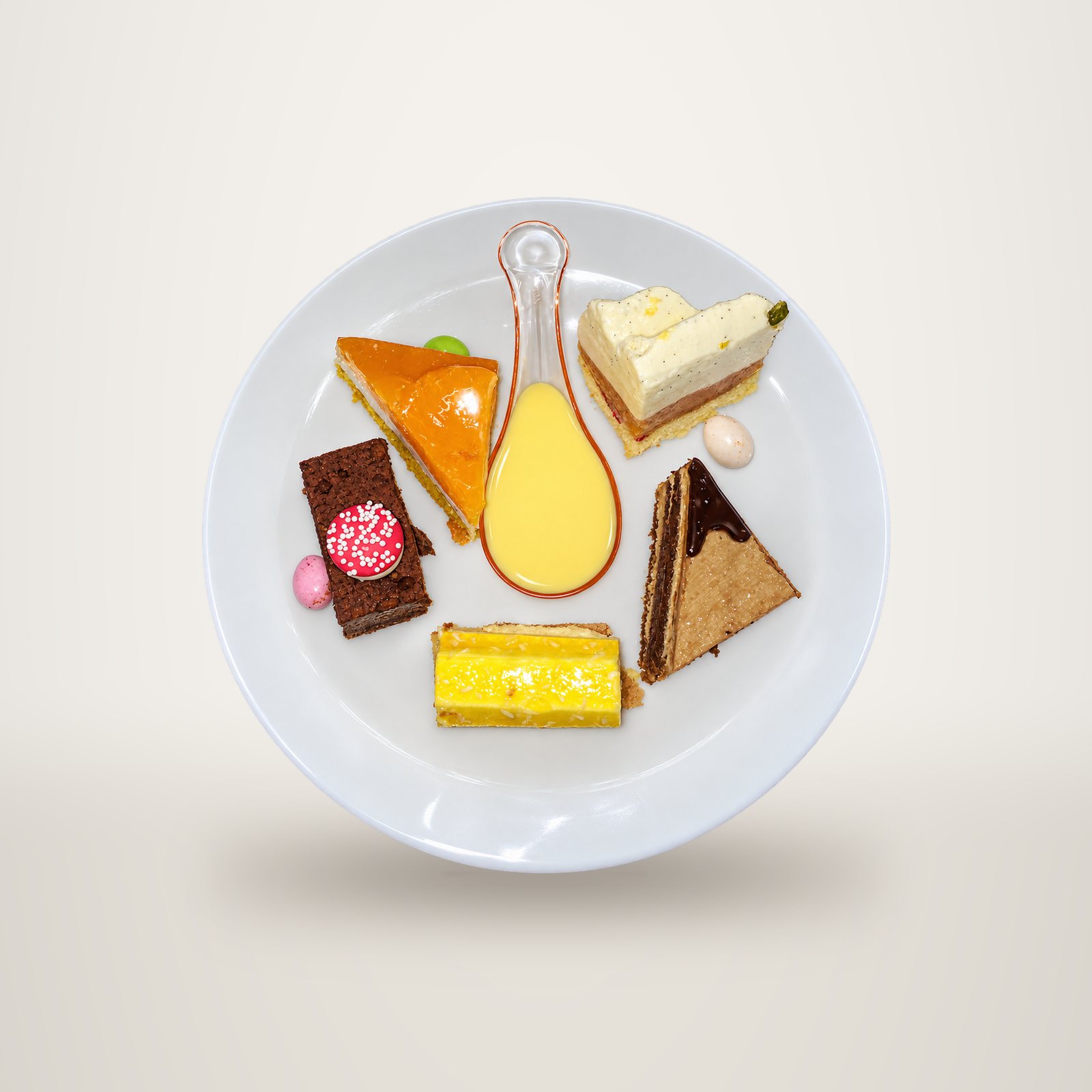 Vue de dessus d'une assiette blanche garnie de cinq petits gâteaux colorés et d'une cuillère de crème anglaise, posée sur un plan de travail en inox.