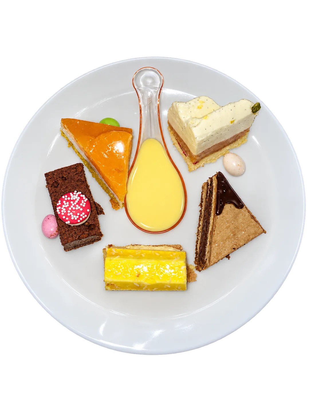 Vue de dessus d'une assiette blanche garnie de cinq petits gâteaux colorés et d'une cuillère de crème anglaise, posée sur un plan de travail en inox.