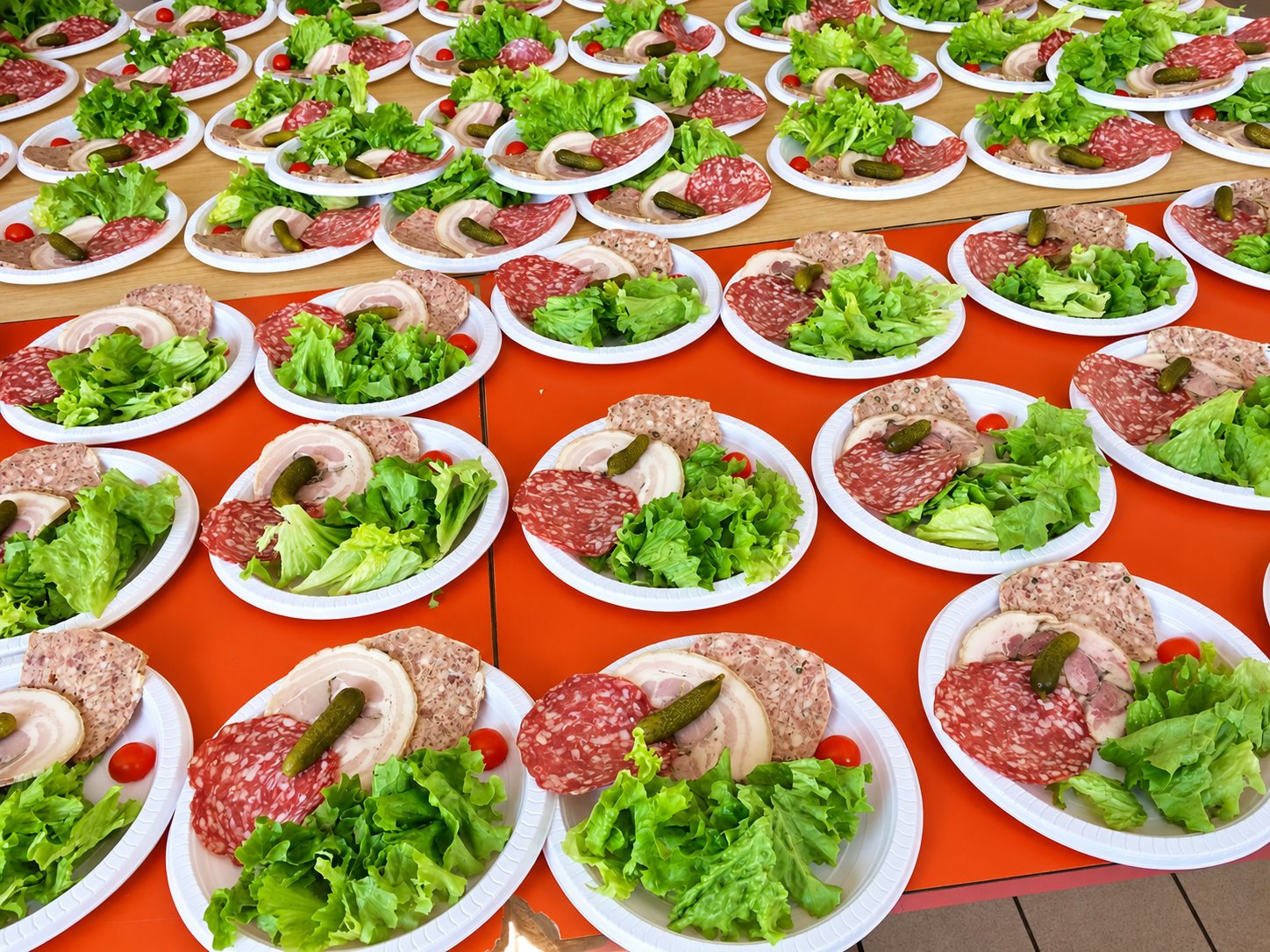De nombreuses assiettes en plastique blanc garnies de saucisson, pâté, lard, cornichons et salade verte sont alignées sur des tables orange.