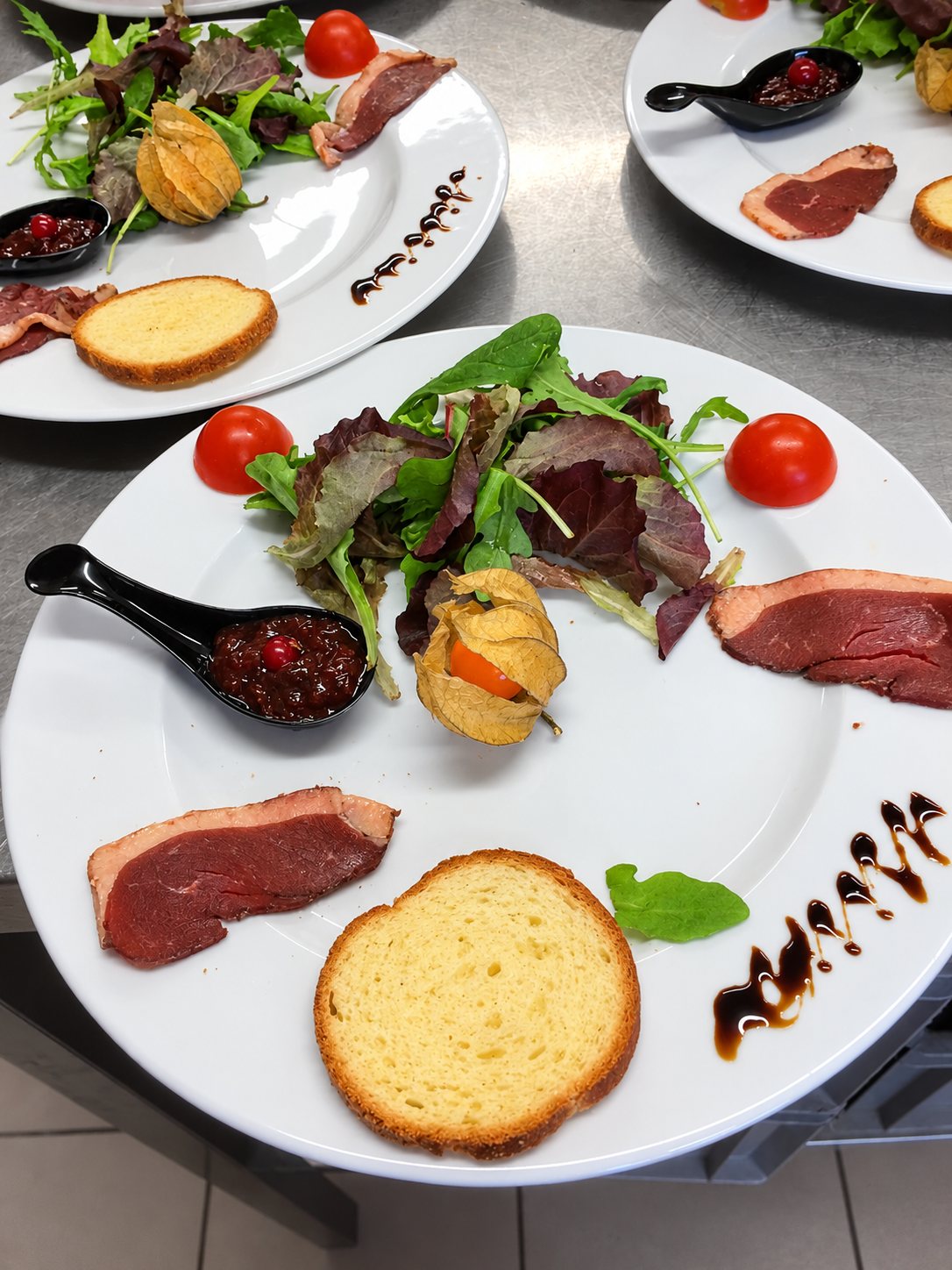 Plusieurs assiettes blanches dressées avec des tranches de magret fumé, de la salade, une physalis, une brioche toastée, des tomates cerises et une cuillère de chutney, posées sur un plan de travail en inox.