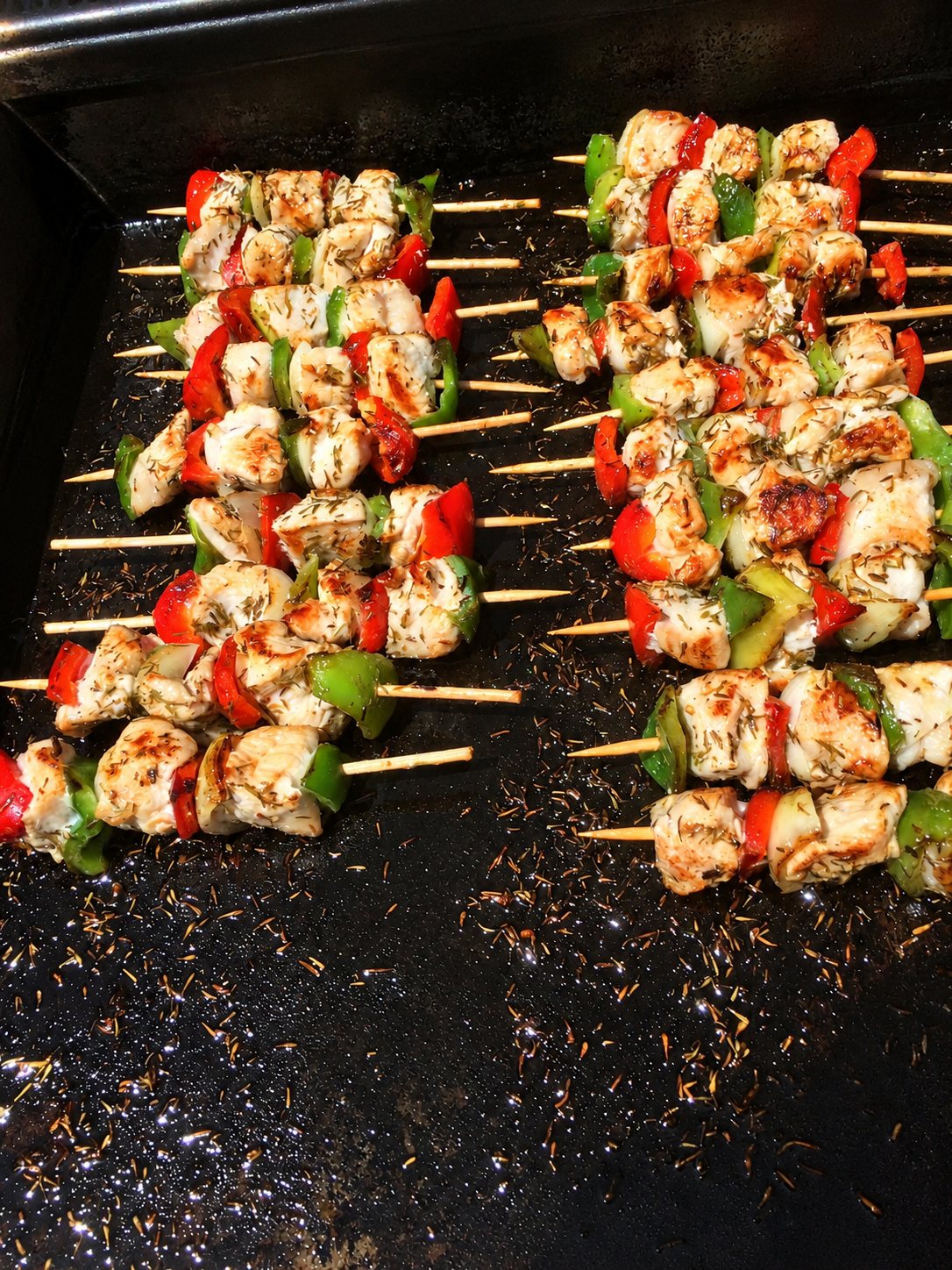 Des brochettes de poulet, de poivrons rouges et verts parsemées de thym cuisent sur une plancha noire.