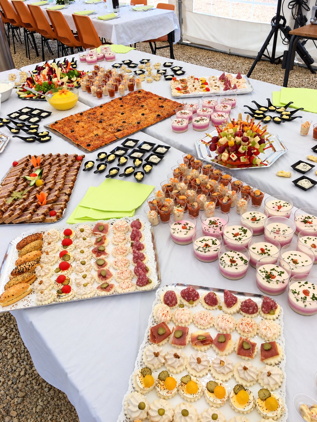 Plusieurs tables recouvertes de nappes blanches présentent un grand buffet de canapés, de verrines, de cuillères apéritives, de pizza découpée et de brochettes de fruits et de fromage, installé sous une tente de réception.