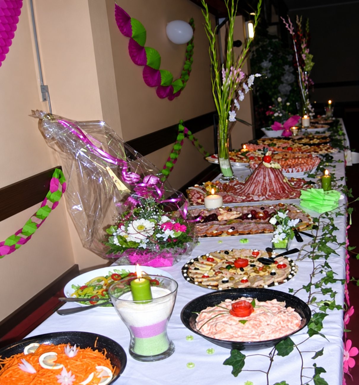 Une longue table de buffet couverte de plats garnis, de bougies et de fleurs, dans une salle décorée de guirlandes roses et vertes.