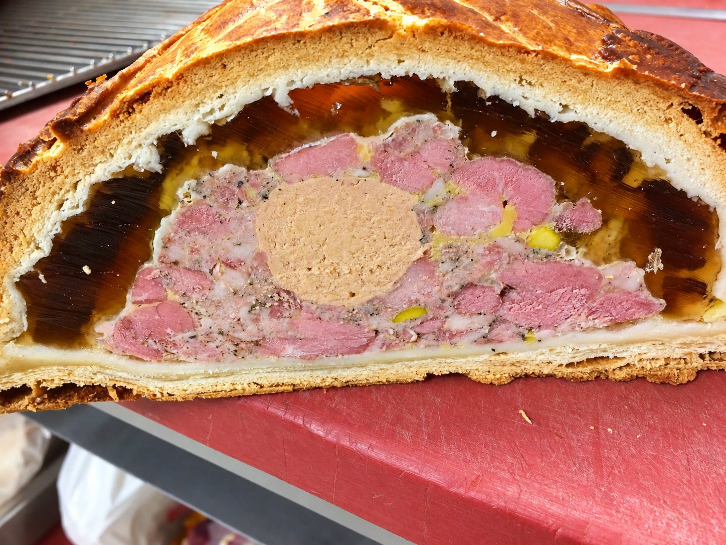 Gros plan sur un pâté en croûte coupé en deux, montrant la viande rose, un centre de foie gras et la gelée ambrée, posé sur un plan de travail rouge.
