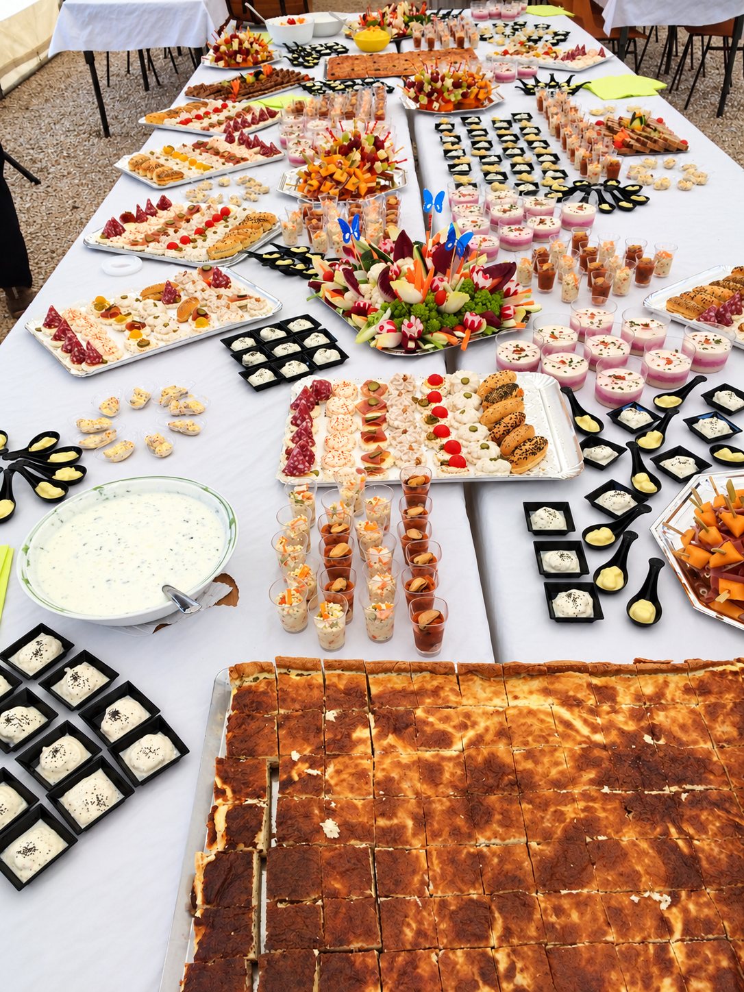 Une longue table couverte d'une nappe blanche présente un grand buffet de canapés, de verrines, de cuillères apéritives et de parts de quiche découpées.
