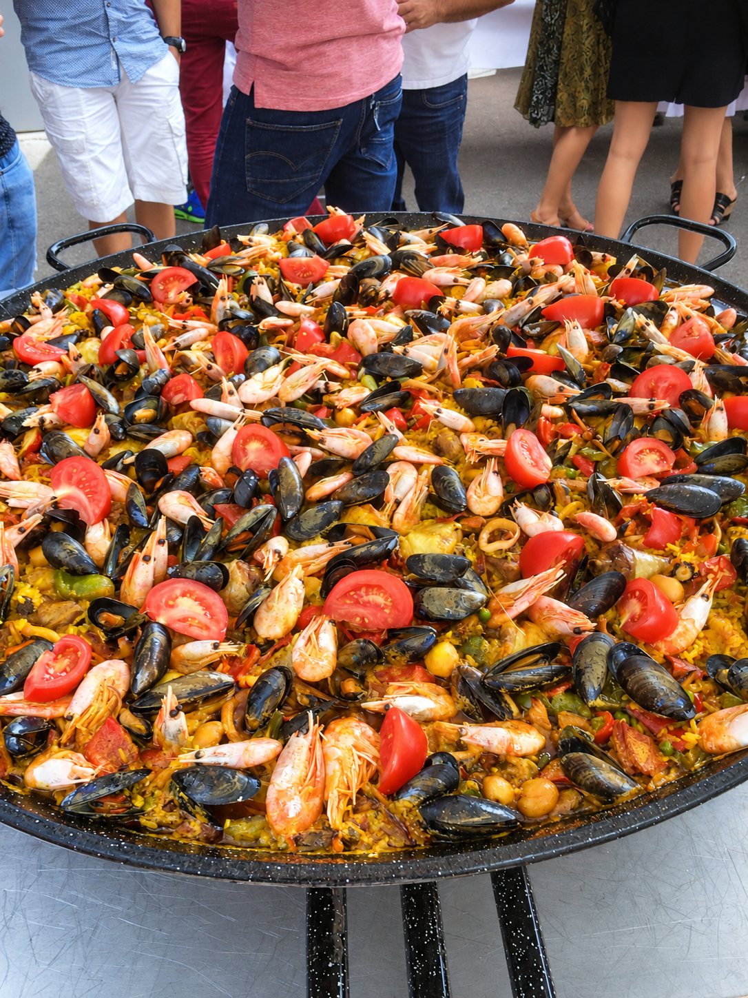Une immense poêle de paëlla garnie de moules, de crevettes et de rondelles de tomate, avec des personnes debout derrière.