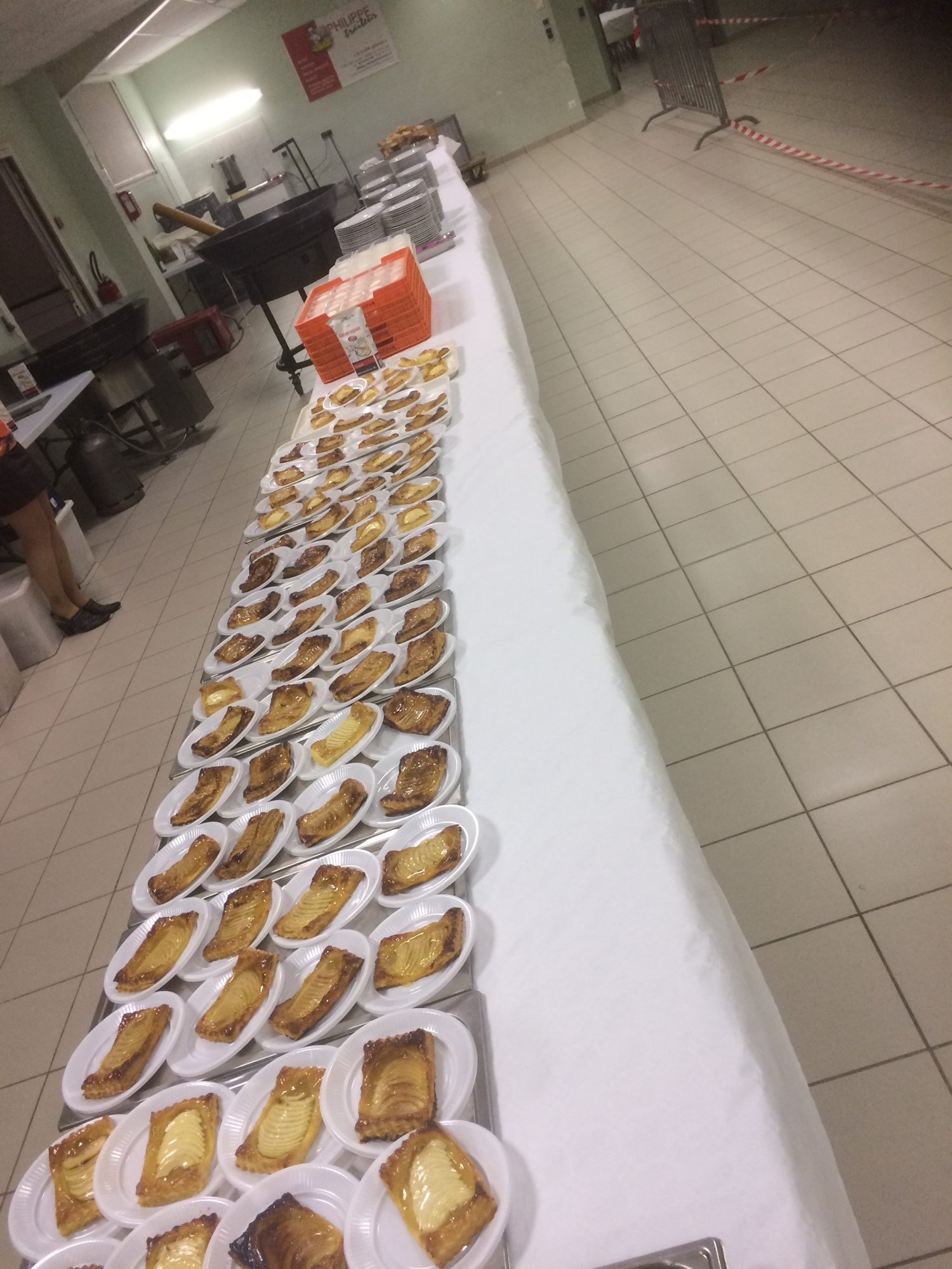 Une très longue table recouverte d'une nappe blanche porte des dizaines d'assiettes blanches garnies chacune d'une tarte feuilletée aux poires, dans une grande salle carrelée.