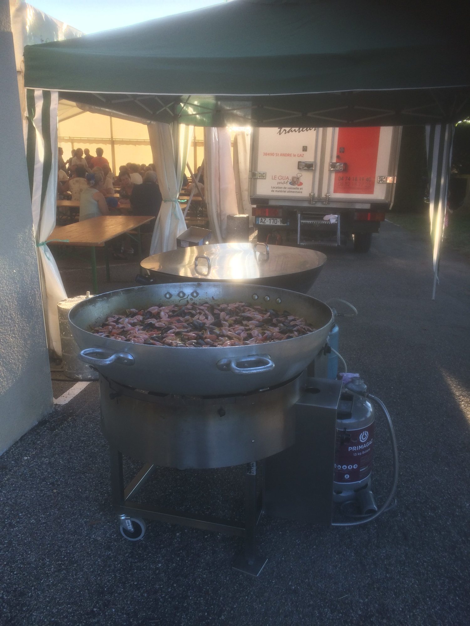 Une immense poêle de paella aux fruits de mer cuit sur un brûleur à gaz sous une tente, avec un camion de traiteur et des convives attablés en arrière-plan.