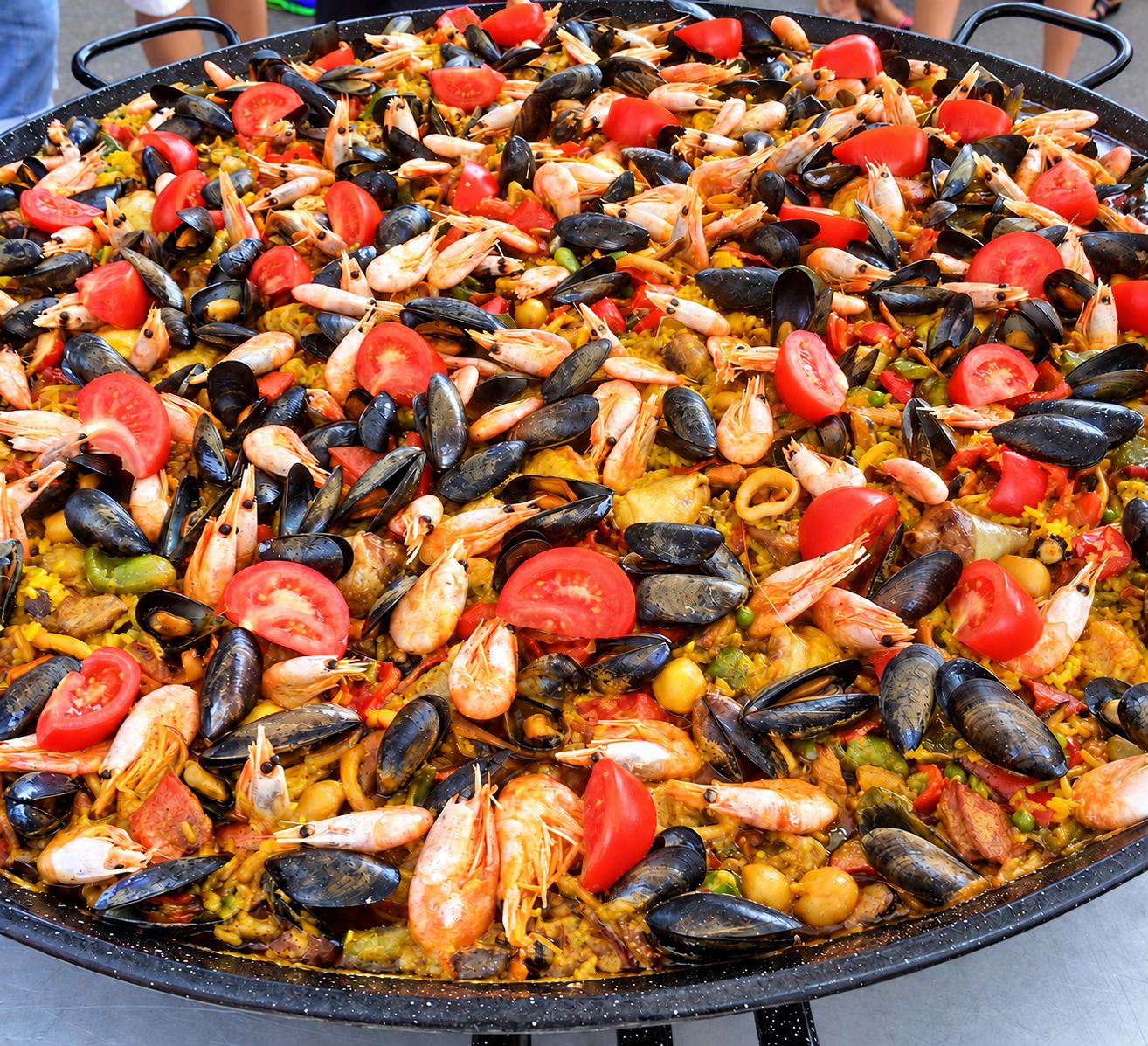 Une grande poêle de paella garnie de moules, de crevettes roses et de rondelles de tomate sur du riz jaune.