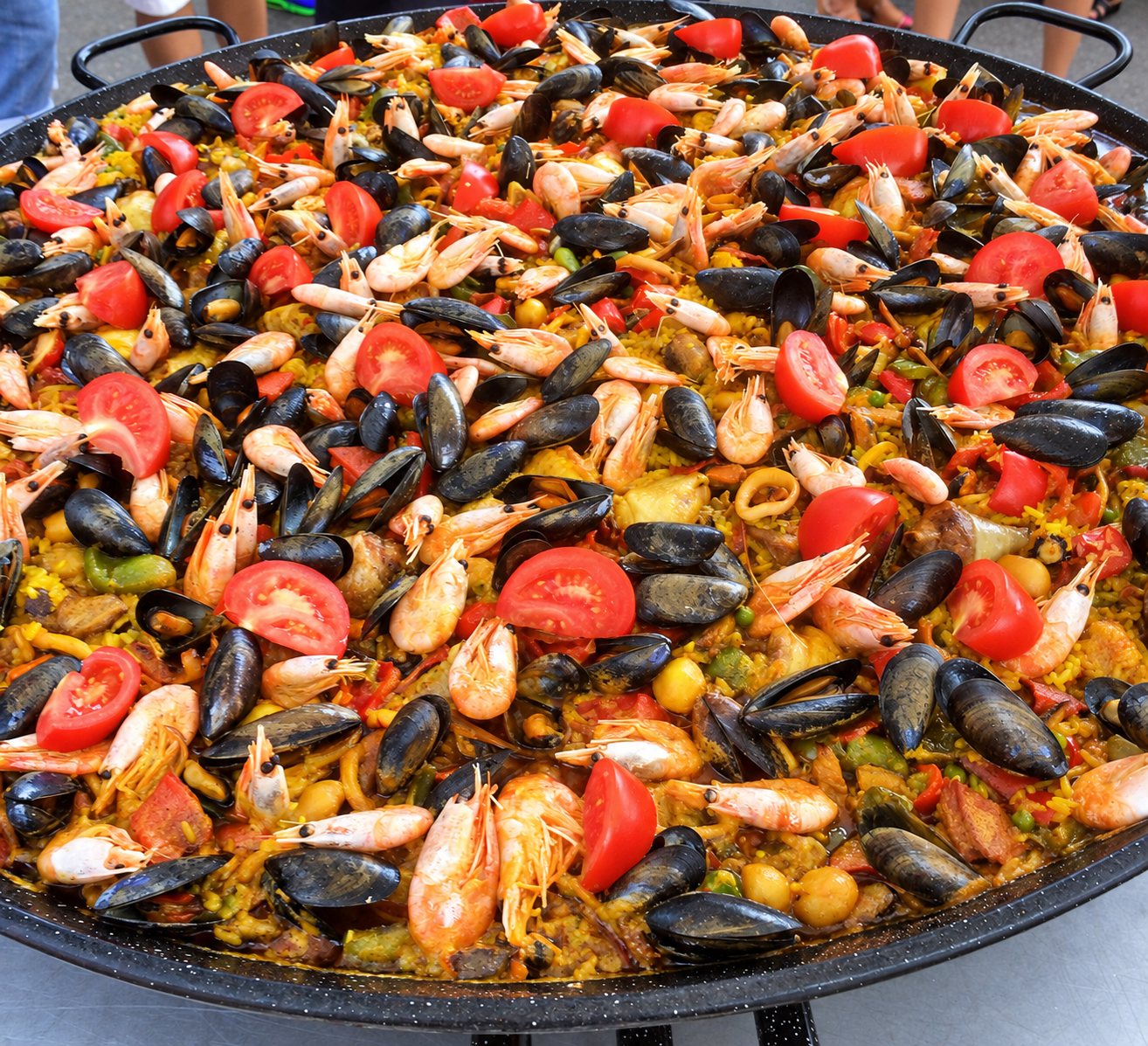 Une grande poêle de paella garnie de moules, de crevettes roses et de rondelles de tomate sur du riz jaune.