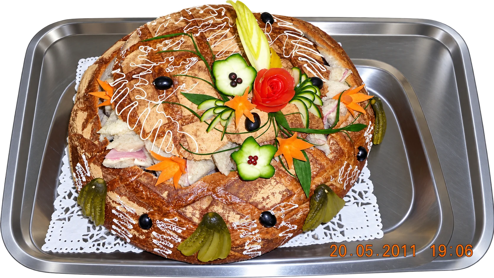 Un gros pain rond garni de sandwichs, décoré de fleurs en concombre, d'une rose de tomate, de carottes, d'olives et de cornichons, posé sur un plateau en inox.