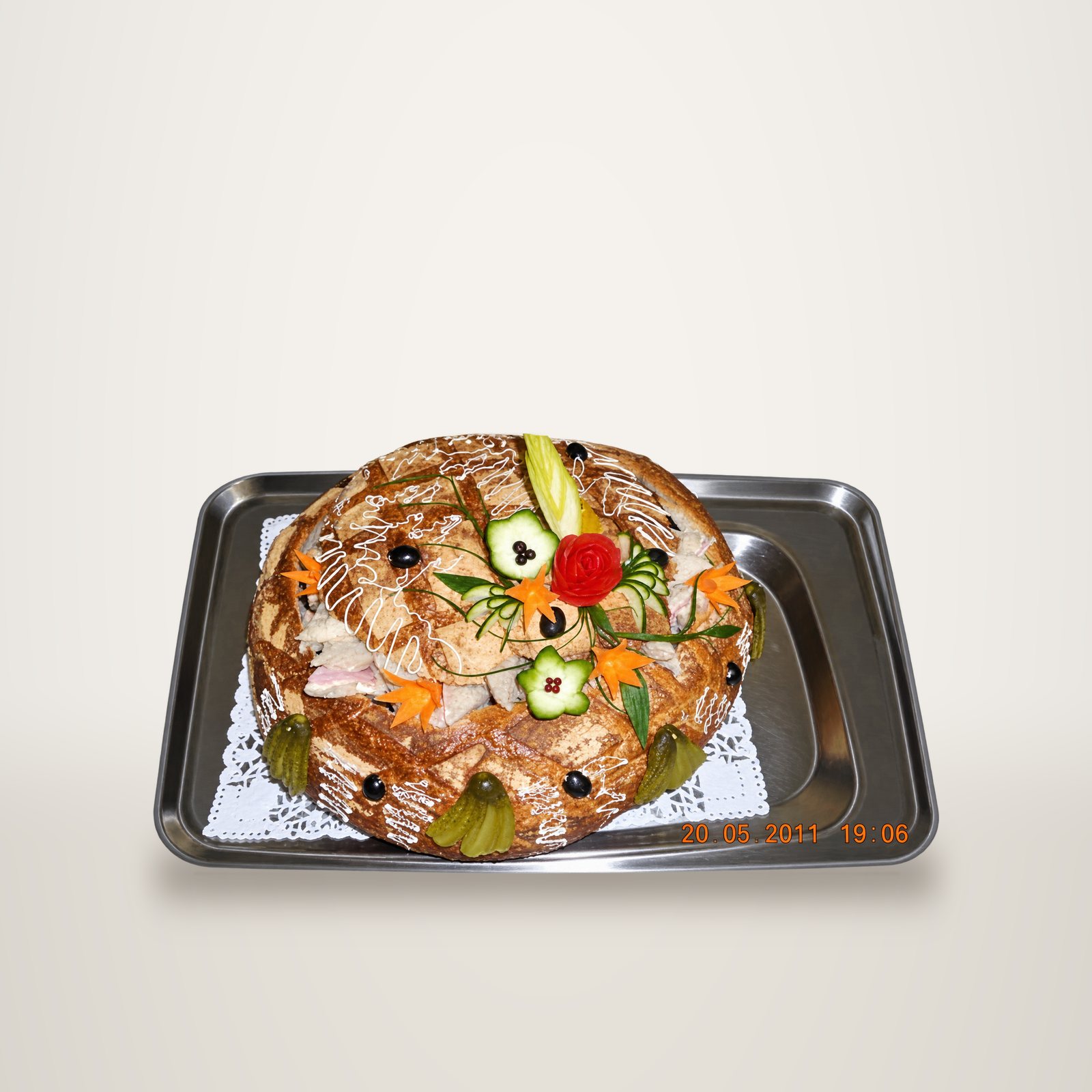 Un grand pain surprise garni de fleurs en légumes sculptés, de cornichons et d'olives, posé sur un plateau en inox avec un napperon en dentelle.