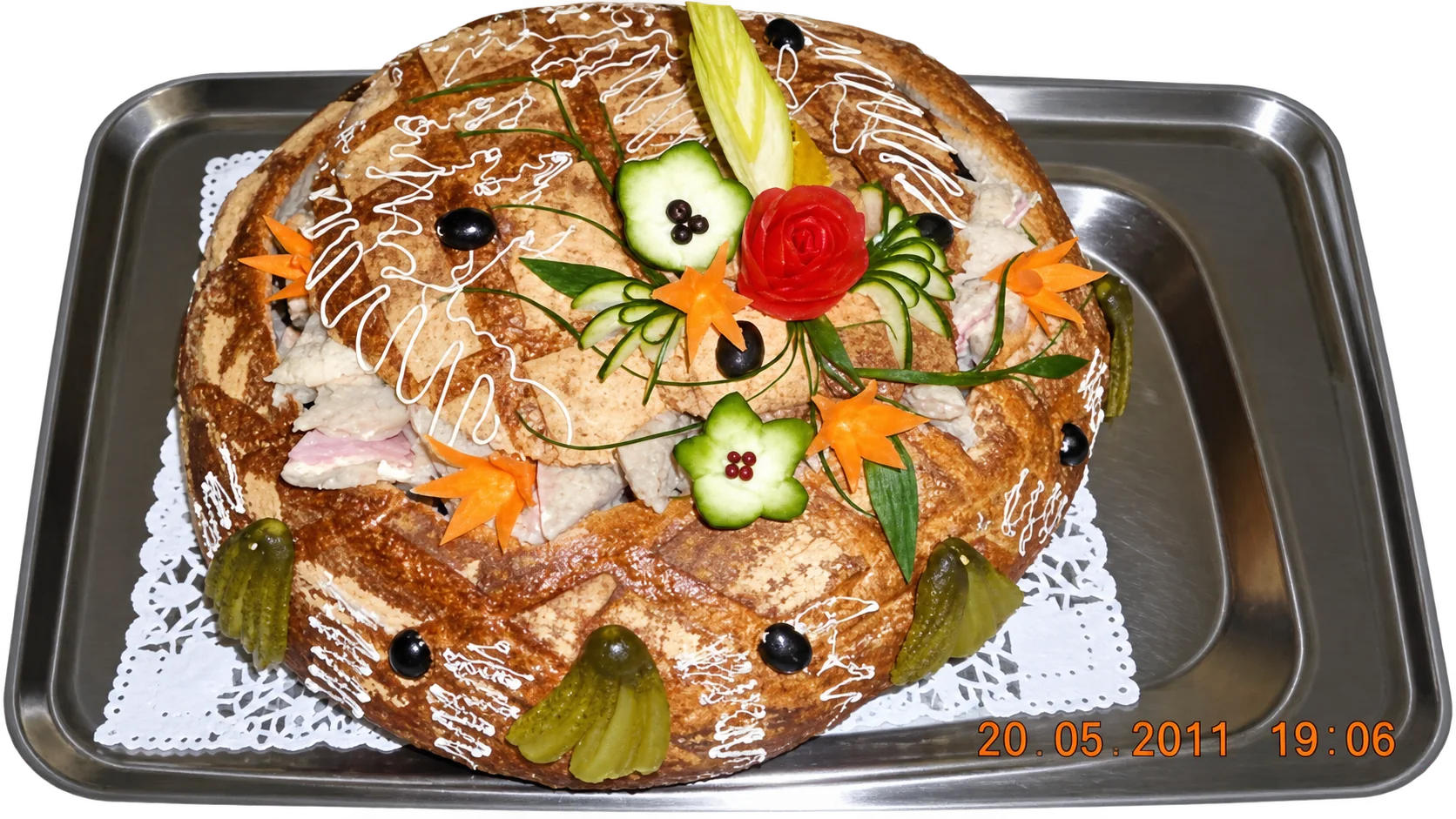 Un grand pain surprise garni de fleurs en légumes sculptés, de cornichons et d'olives, posé sur un plateau en inox avec un napperon en dentelle.