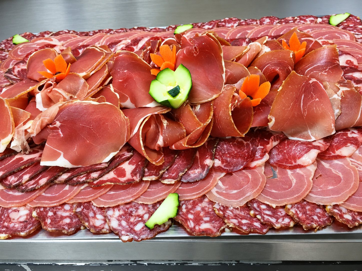 Un grand plateau métallique garni de tranches de jambon cru, de coppa, de pancetta et de saucisson, décoré de fleurs de carotte et de courgette.