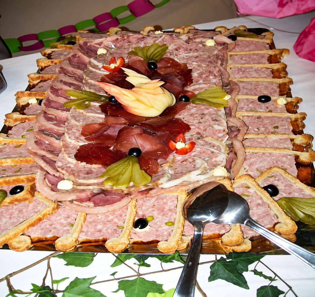 Un grand plateau de pâté en croûte en tranches, garni d'olives noires, de cornichons, de jambon cru et d'une décoration sculptée, posé sur une table de buffet avec deux cuillères de service.