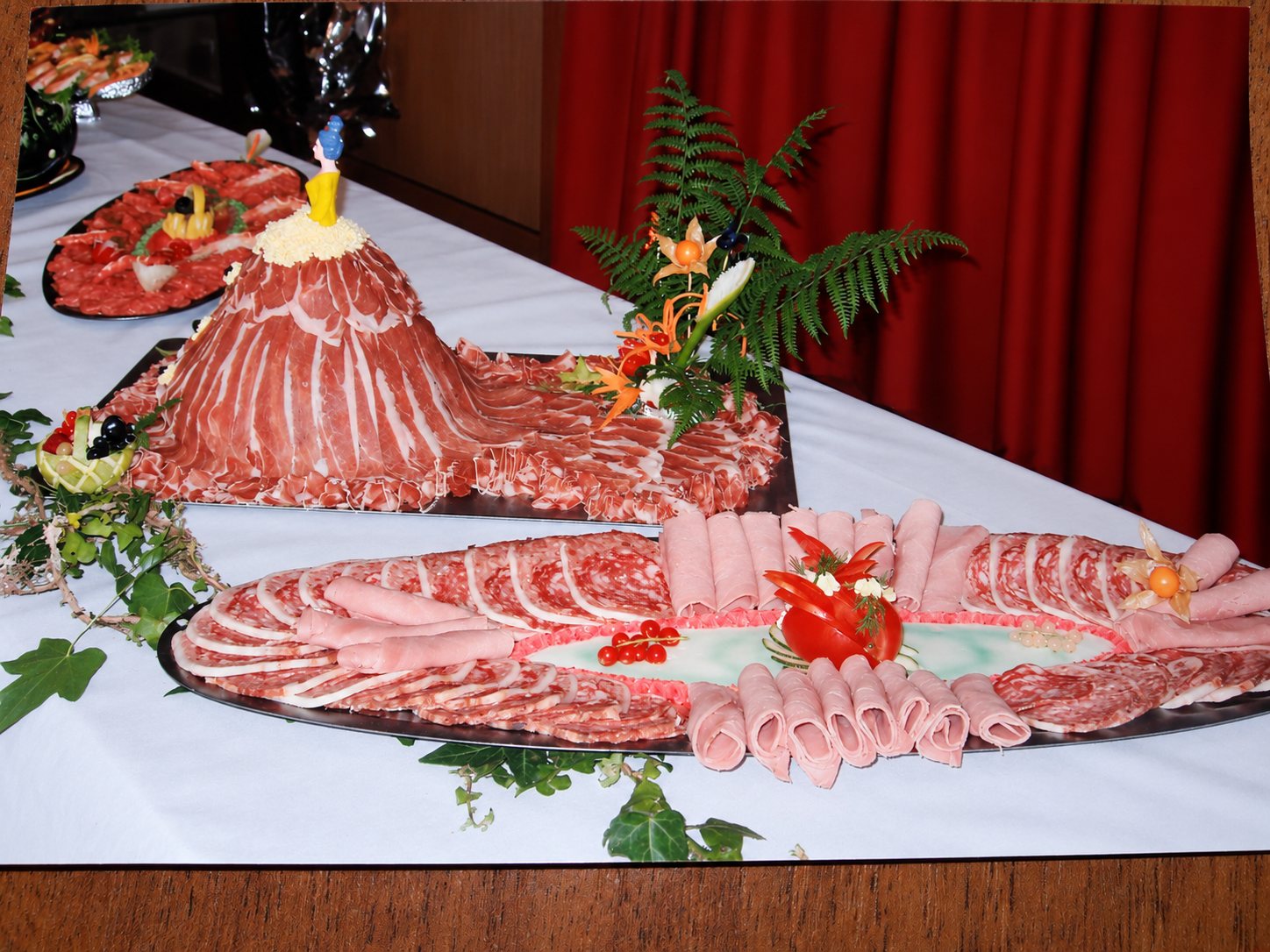 Sur une table recouverte d'une nappe blanche, des plateaux de charcuterie en tranches et roulées sont décorés de tomate, de fougères et d'une figurine en forme de robe de jambon.