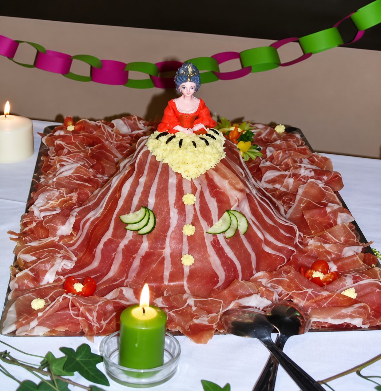 Une poupée en corsage rouge dont la longue robe est faite de tranches de jambon cru décorées de concombre, posée sur une table de buffet avec des bougies.