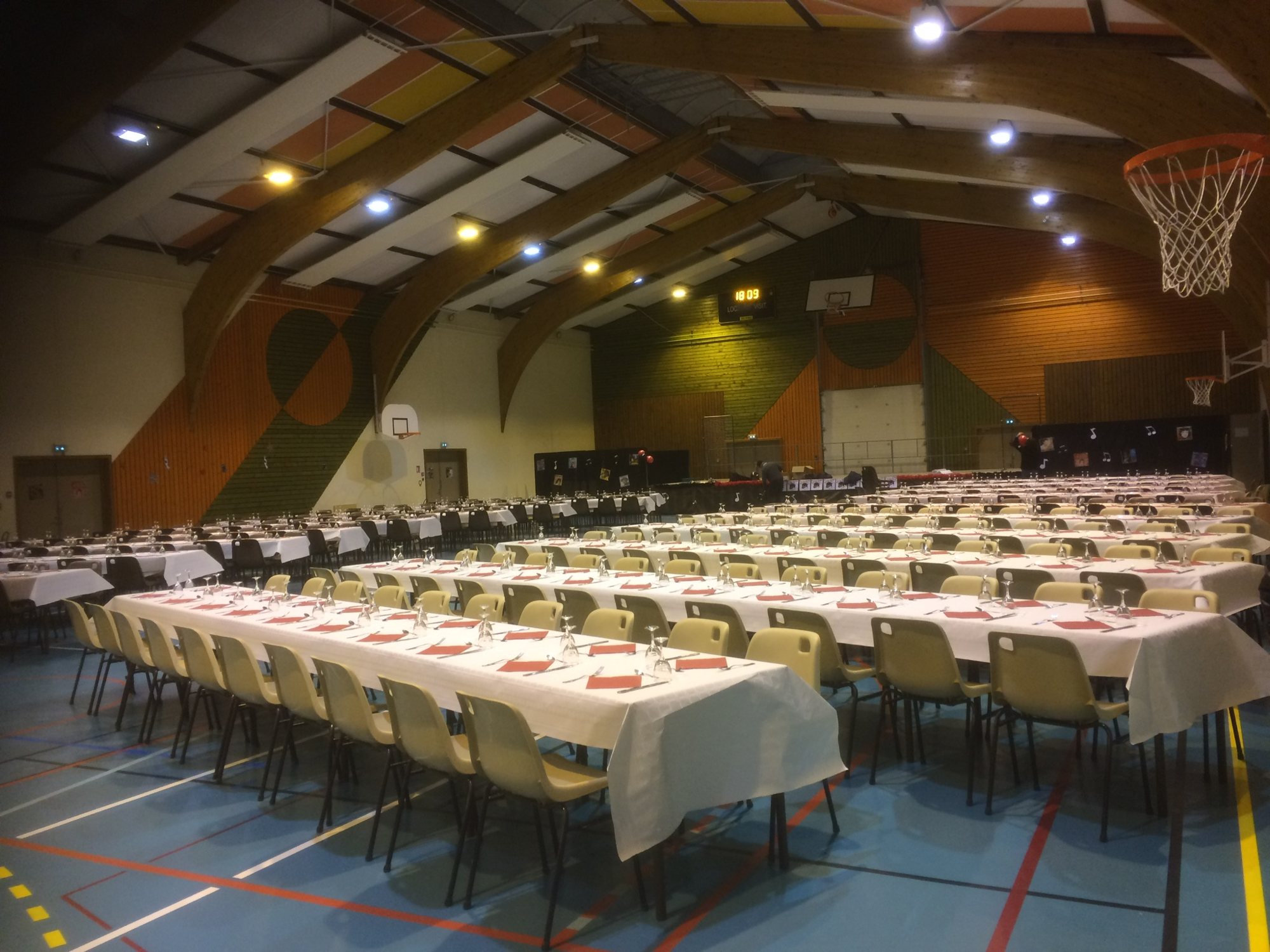 Un gymnase aux poutres de bois cintrées est rempli de longues tables à nappes blanches, de chaises beiges et de verres, prêt pour un grand repas.