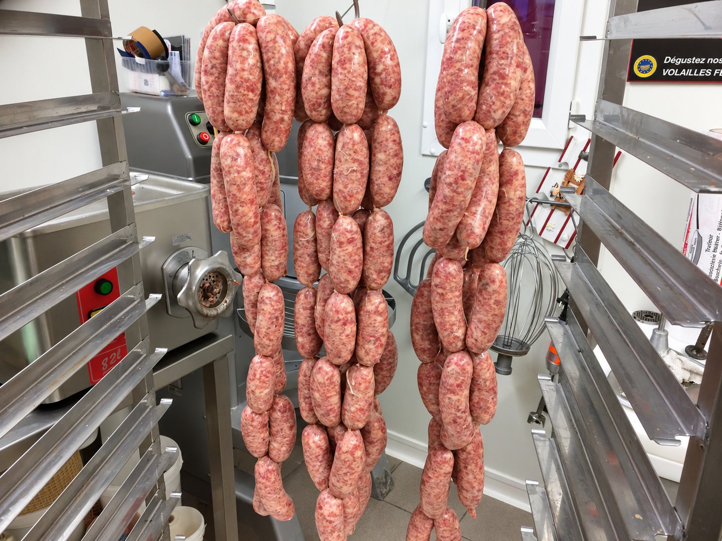 Trois longs chapelets de saucisses fraîches pendent dans l'atelier d'un boucher, entre des étagères en inox et un hachoir à viande.