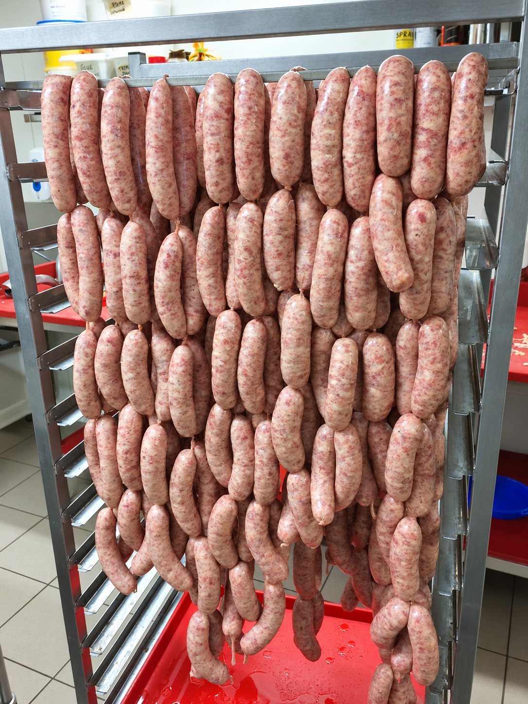 De nombreuses saucisses crues sont accrochées en chapelets sur un chariot en inox, au-dessus d'un bac rouge, dans un laboratoire de boucherie.