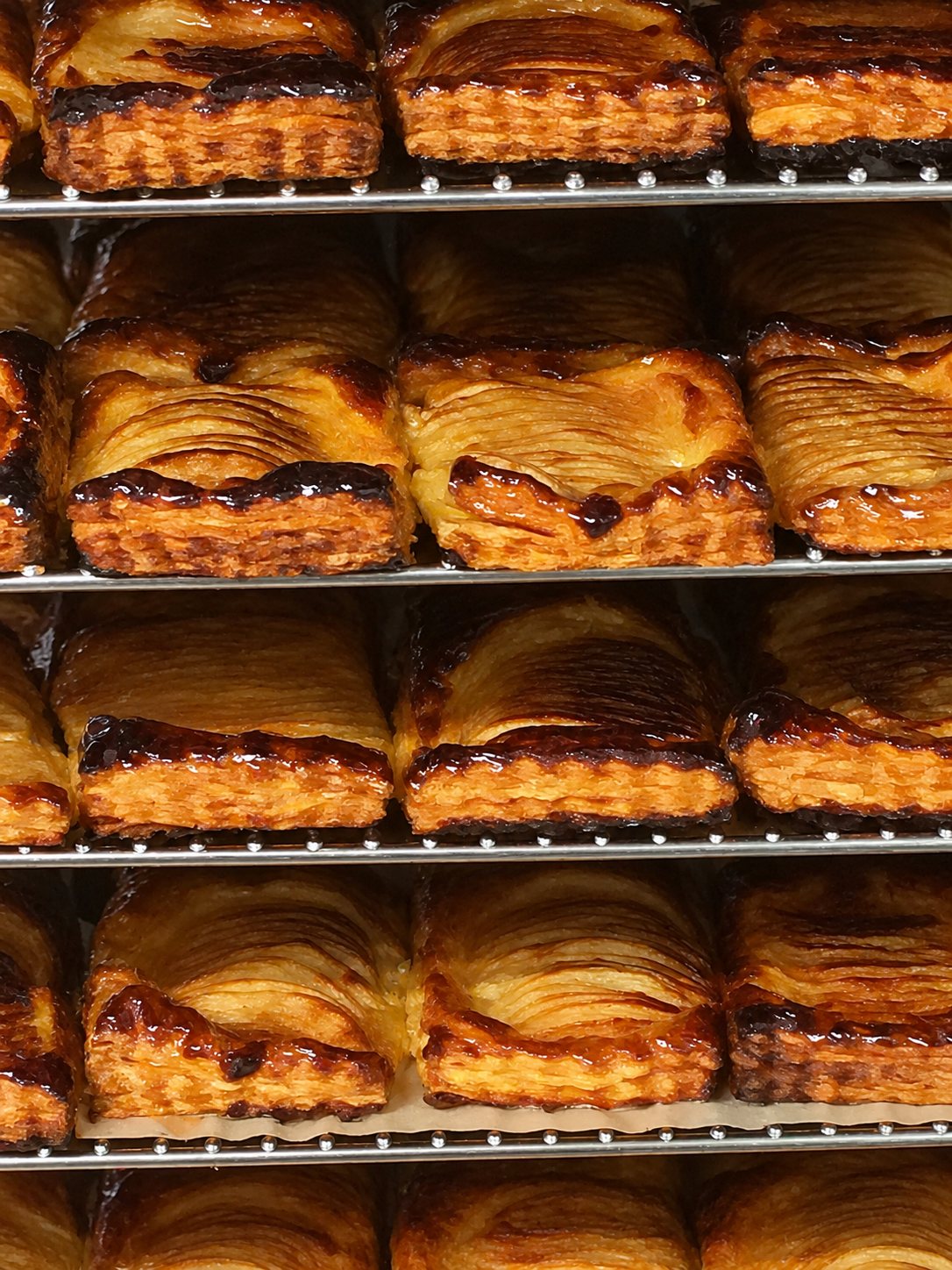 Plusieurs étagères métalliques supportent des rangées de viennoiseries feuilletées caramélisées, fraîchement sorties du four.